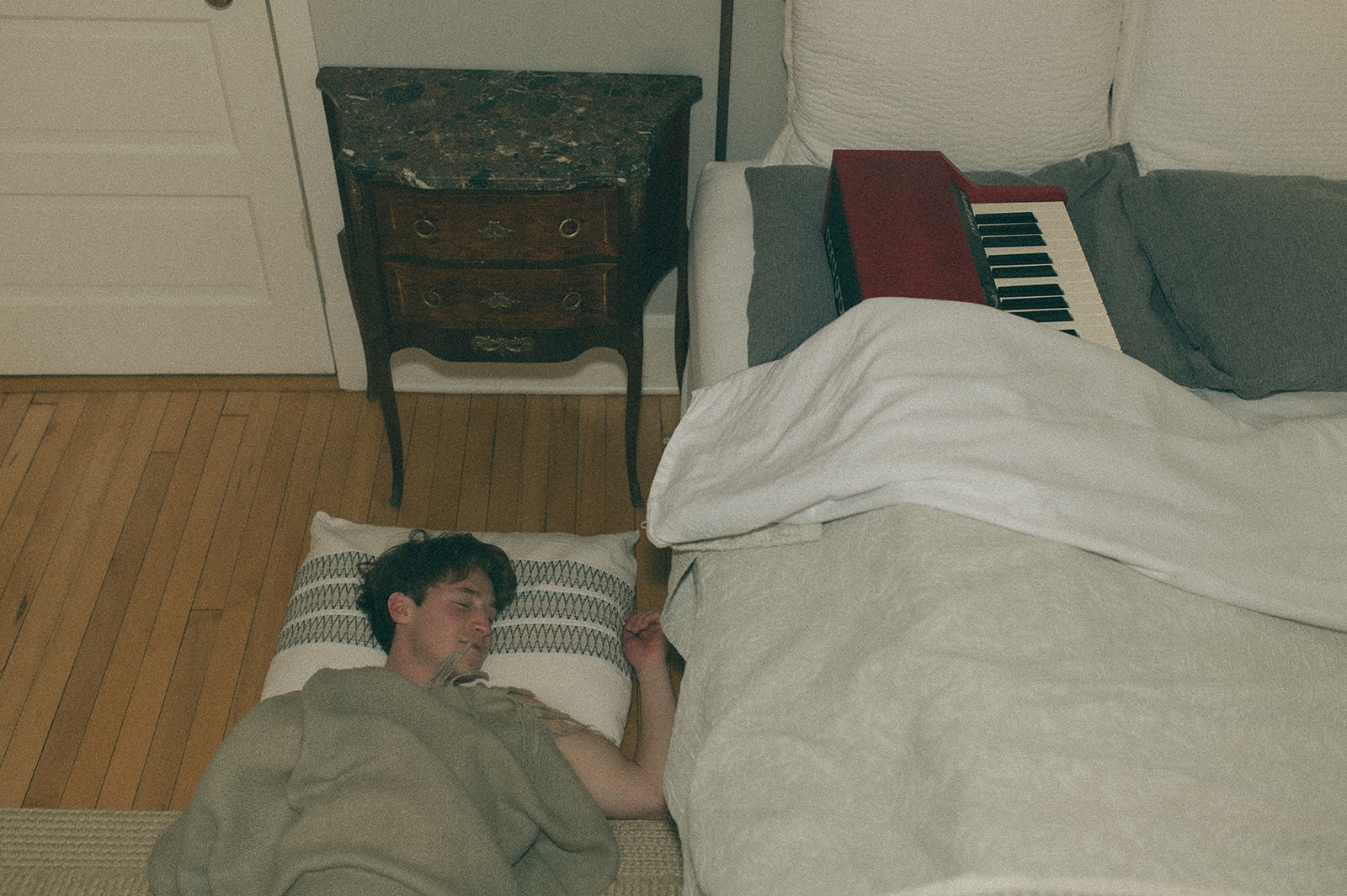 Cam asleep on the floor beside a bed with a small red piano on it